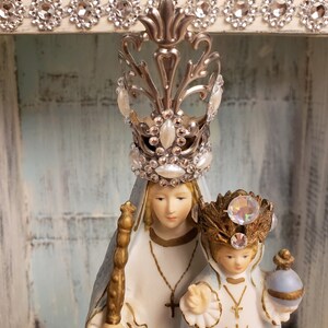 May include: A porcelain figurine of the Virgin Mary and baby Jesus. The Virgin Mary is wearing a blue and white robe and a silver crown with rhinestones. The baby Jesus is wearing a white robe and a gold crown with rhinestones. The figurine is displayed in a white wooden frame.