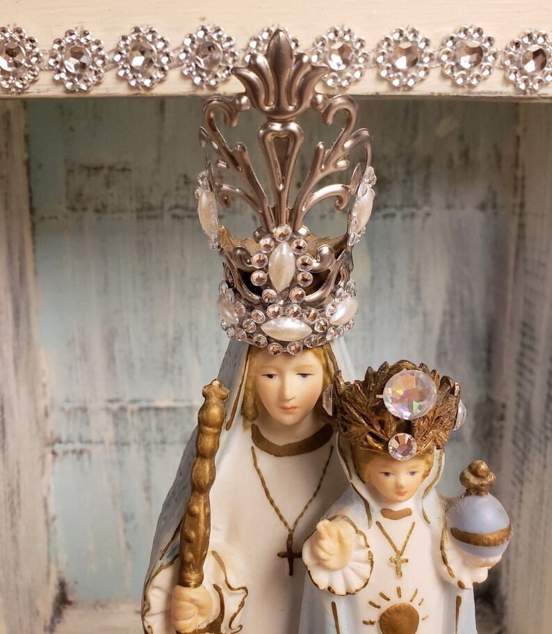 May include: A porcelain figurine of the Virgin Mary and baby Jesus. The Virgin Mary is wearing a blue and white robe and a silver crown with rhinestones. The baby Jesus is wearing a white robe and a gold crown with rhinestones. The figurine is displayed in a white wooden frame.