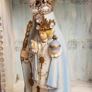 May include: A white porcelain figurine of the Virgin Mary, wearing a blue robe and a silver crown with rhinestones. She is holding a golden staff and a small globe in her hands.