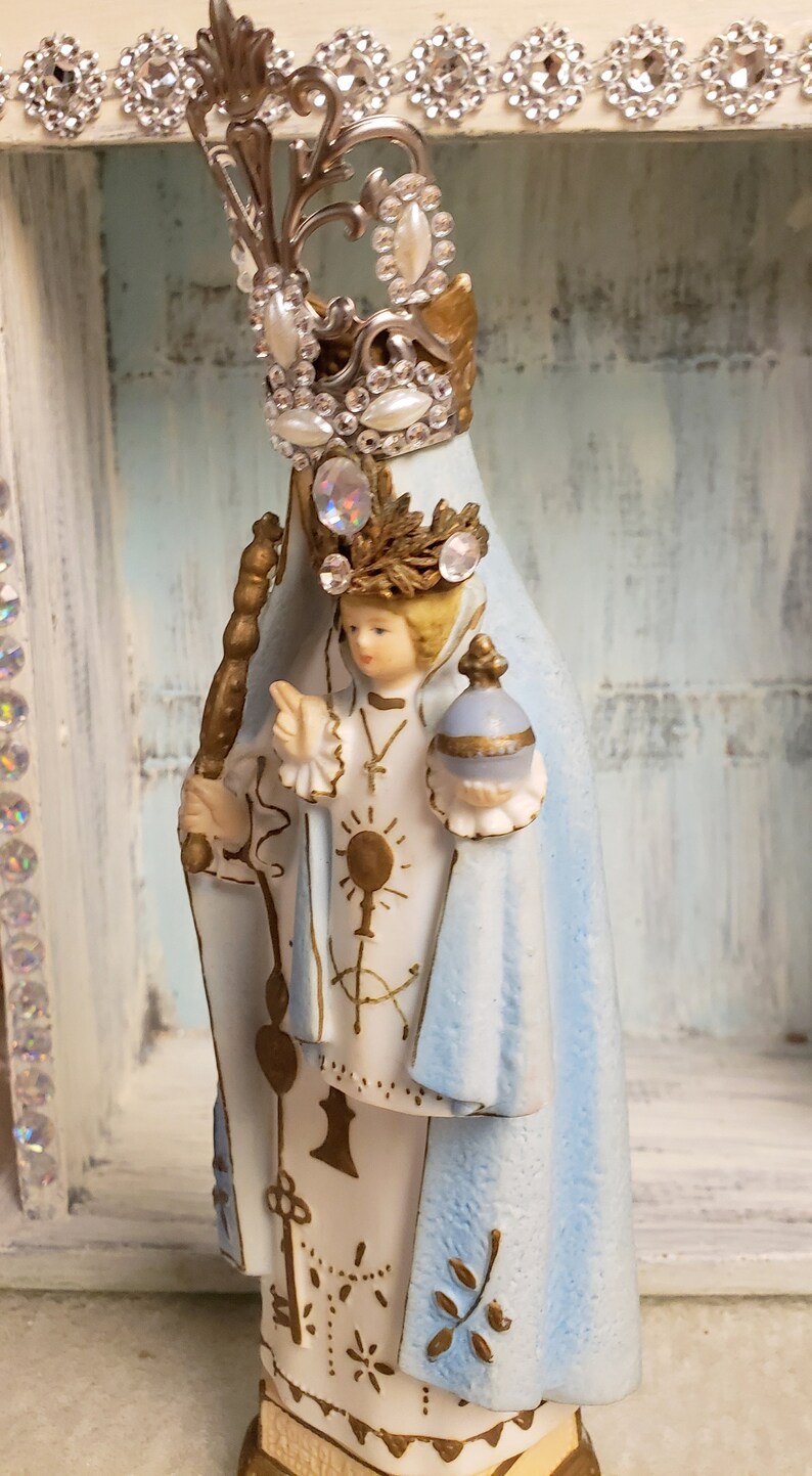 May include: A white porcelain figurine of the Virgin Mary, wearing a blue robe and a silver crown with rhinestones. She is holding a golden staff and a small globe in her hands.
