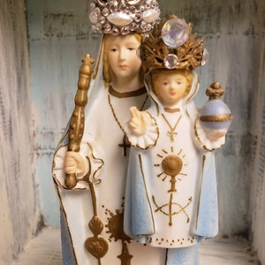 May include: A white porcelain figurine of the Virgin Mary and baby Jesus. Mary is wearing a blue robe with gold accents and a crown with white pearls and rhinestones. Jesus is wearing a blue robe with gold accents and a crown with rhinestones. The figurine is standing on a wooden surface.