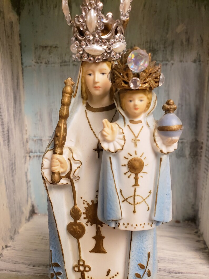 May include: A white porcelain figurine of the Virgin Mary and baby Jesus. Mary is wearing a blue robe with gold accents and a crown with white pearls and rhinestones. Jesus is wearing a blue robe with gold accents and a crown with rhinestones. The figurine is standing on a wooden surface.