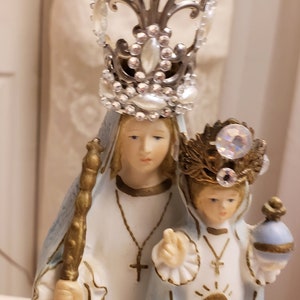 May include: A white porcelain figurine of the Virgin Mary and baby Jesus. The Virgin Mary is wearing a blue robe and a silver crown with rhinestones. The baby Jesus is wearing a blue robe and a gold crown with rhinestones. The figurine is a religious icon.