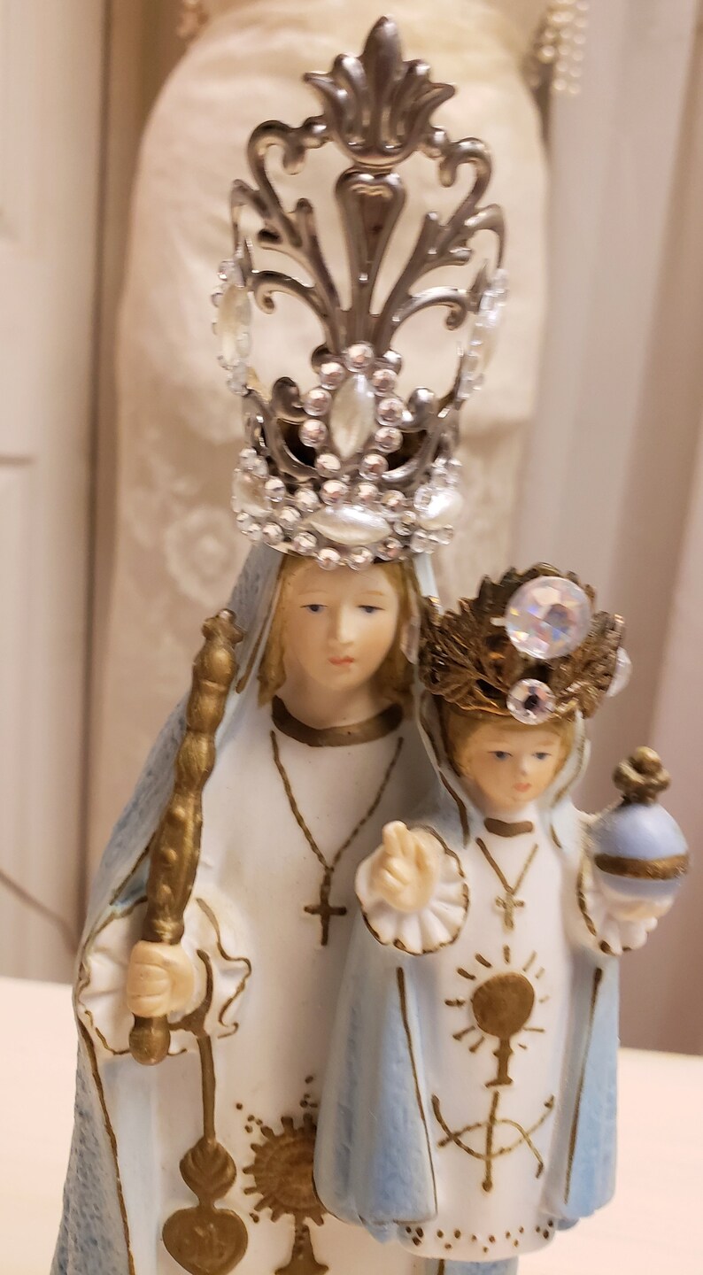 May include: A white porcelain figurine of the Virgin Mary and baby Jesus. The Virgin Mary is wearing a blue robe and a silver crown with rhinestones. The baby Jesus is wearing a blue robe and a gold crown with rhinestones. The figurine is a religious icon.