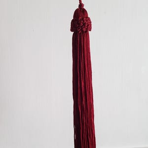 May include: A long, burgundy tassel with a decorative, rounded top. The tassel is suspended from a metal rod against a white background. The tassel's strands are neatly aligned, creating a visually appealing, decorative element.