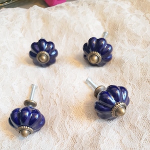 May include: Four navy blue ceramic cabinet knobs with a scalloped design and brass-colored hardware. The knobs are arranged on a white lace surface, showcasing their decorative appeal. Each knob has a stem for easy installation.