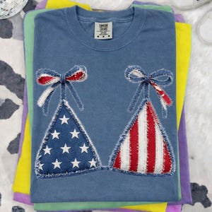 Könnte beinhalten: Ein blaues Comfort Colors T-Shirt mit einem patriotischen Bikini-Design. Eine Seite hat ein denimartiges Dreieck mit weißen Sternen, die andere rote und weiße Streifen. Beide sind mit roten, weißen und blauen Schleifen verziert. Das T-Shirt ist mit anderen farbigen Shirts gefaltet.