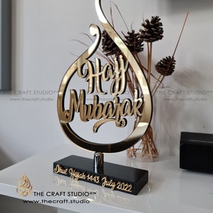 Peut inclure: Un signe décoratif doré "Hajj Mubarak" avec une écriture élégante, monté sur une base noire. La base porte l'inscription "Dhul Hijjah 1443 July 2022". Le logo The Craft Studio est visible. Un objet décoratif.