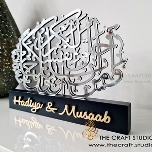 Op de afbeelding: Een zilveren metalen wandkunst met Arabische kalligrafie op een zwarte houten basis. De basis heeft de namen "Hadiya & Musaab" in gouden letters.