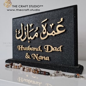 Peut inclure: Plaque noire textur&eacute;e avec paillettes et lettrage dor&eacute;. Le texte arabe "Umrah Mubarak" est en haut, suivi de "Husband, Dad & Nana". Un chapelet est devant. Le logo et le site web de The Craft Studio sont visibles.