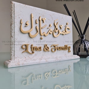 Peut inclure: Plaque d&eacute;corative blanche effet marbre avec lettrage dor&eacute;. La plaque pr&eacute;sente de la calligraphie arabe et le texte anglais "Lina & Family". Le logo et le site web de The Craft Studio sont visibles.