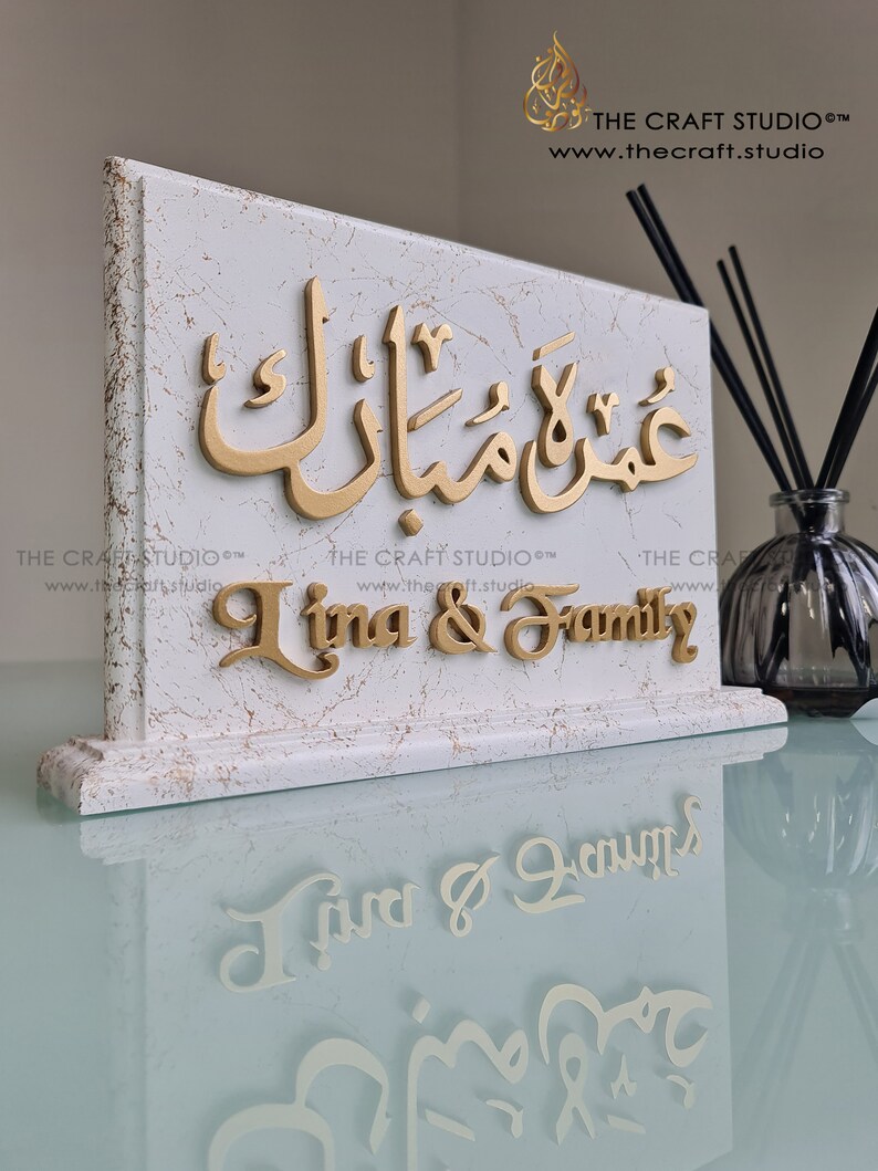 Peut inclure: Plaque d&eacute;corative blanche effet marbre avec lettrage dor&eacute;. La plaque pr&eacute;sente de la calligraphie arabe et le texte anglais "Lina & Family". Le logo et le site web de The Craft Studio sont visibles.