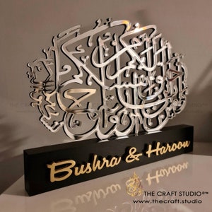 Op de afbeelding: Een zilveren gespiegelde kalligrafie kunstwerk met de namen "Bushra & Haroon" op een zwarte houten basis. De kalligrafie is in Arabisch schrift.