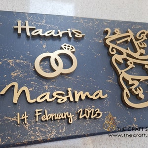May include: A dark grey wooden sign with gold lettering. The sign reads "Haaris & Nasima" with a gold ring design and the date "14 February 2023". The sign also features a gold Arabic calligraphy design.