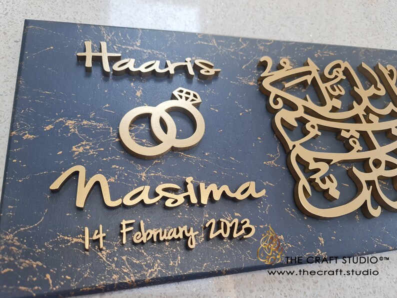 May include: A dark grey wooden sign with gold lettering. The sign reads "Haaris & Nasima" with a gold ring design and the date "14 February 2023". The sign also features a gold Arabic calligraphy design.