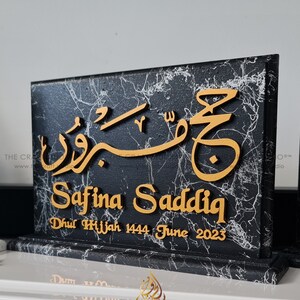 Peut inclure: Plaque noire avec effet marbre et calligraphie arabe dorée. Le nom "Safina Saddiq" est inscrit, ainsi que "Dhul Hijjah 1444 June 2023". La plaque est posée sur une surface blanche.