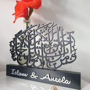 May include: Black wooden sign with intricate Arabic calligraphy and the names "Islam & Aneela" in white lettering. The sign is on a white surface with a vase of red flowers.