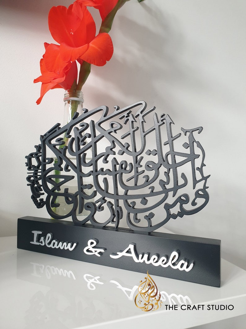 May include: Black wooden sign with intricate Arabic calligraphy and the names "Islam & Aneela" in white lettering. The sign is on a white surface with a vase of red flowers.