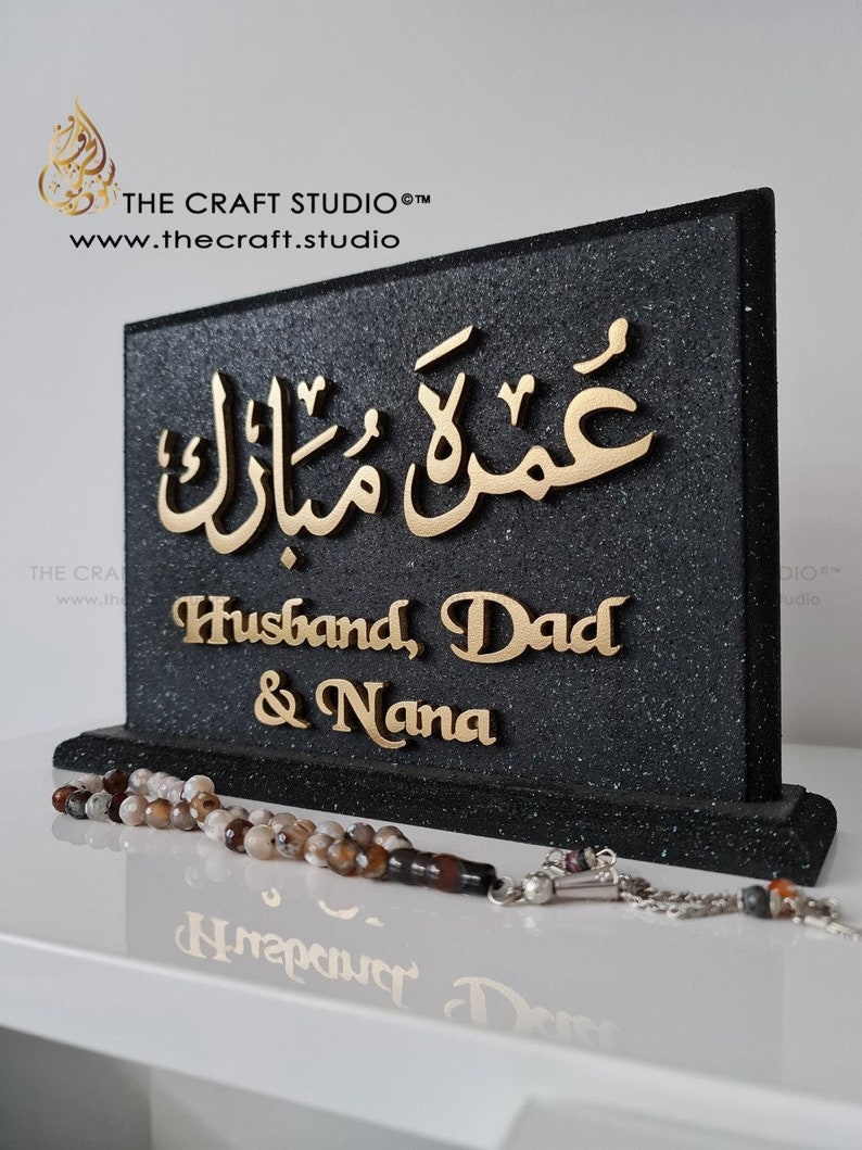 Peut inclure: Plaque noire textur&eacute;e avec paillettes et lettrage dor&eacute;. Le texte arabe "Umrah Mubarak" est en haut, suivi de "Husband, Dad & Nana". Un chapelet est devant. Le logo et le site web de The Craft Studio sont visibles.