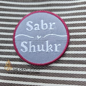 Puede incluir: Un parche redondo bordado gris con las palabras "Sabr Shukr" en escritura blanca. El parche tiene un borde granate y está sobre un fondo a rayas. El texto está separado por un pequeño elemento decorativo.