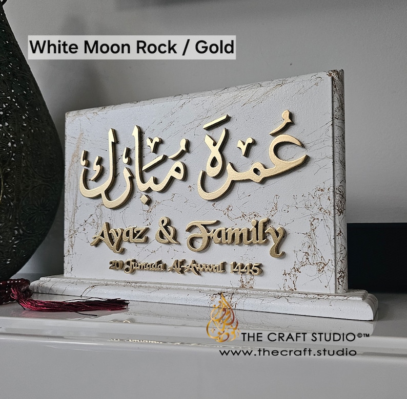 Peut inclure: Plaque d&eacute;corative blanche et dor&eacute;e avec calligraphie arabe et le texte "Ayaz & Family". La plaque a une texture marbr&eacute;e et est inscrite d'une date. Le texte "White Moon Rock / Gold" est en haut.