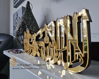 May include: A decorative gold and black wall art piece featuring Arabic calligraphy. The artwork is made of layered materials, with a reflective gold surface and black backing. The Craft Studio logo is visible. The piece is displayed on a white surface.