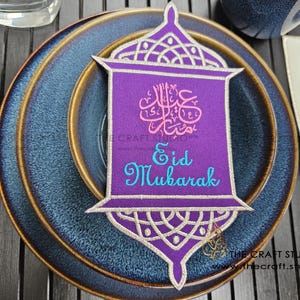 Puede incluir: Decoración bordada morada de Eid Mubarak con forma de linterna, con detalles dorados y blancos. La decoración está colocada sobre un plato azul con borde dorado. El texto "Eid Mubarak" está escrito en azul.