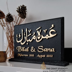 Peut inclure: Panneau en bois noir avec lettrage dor&eacute;. Le panneau pr&eacute;sente une &eacute;criture arabe et les noms "Bilal & Sana" ainsi que la date "ao&ucirc;t 2022". Le panneau est un objet d&eacute;coratif, peut-&ecirc;tre pour un mariage ou une occasion sp&eacute;ciale.