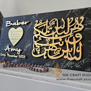 May include: A black and gold wooden plaque with the names "Baber" and "Amy" and the date "24th November 2023". The plaque also features a gold Arabic calligraphy design and a small heart-shaped plaque with a quote about love.