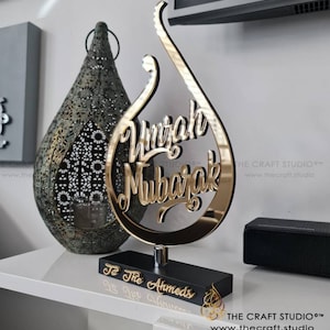 Peut inclure: Un signe doré "Umrah Mubarak" sur une base noire, avec le texte "To The Ahmeds". Le signe a une forme de goutte d'eau stylisée. Une lanterne décorative et un haut-parleur sont en arrière-plan. Le logo The Craft Studio est visible.