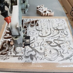 May include: A close-up of a CNC machine cutting intricate Arabic calligraphy designs into a piece of wood. The machine is cutting out the letters of the phrase "The Craft Studio" in a decorative font.