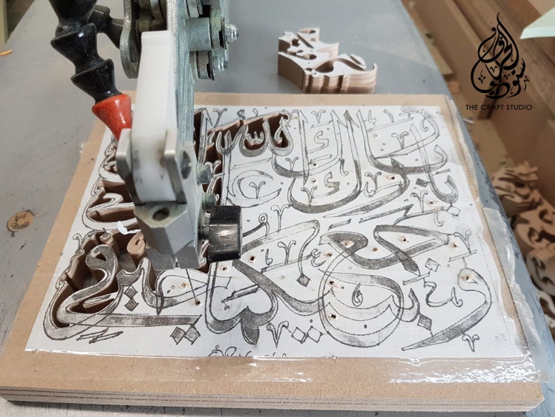 May include: A close-up of a CNC machine cutting intricate Arabic calligraphy designs into a piece of wood. The machine is cutting out the letters of the phrase "The Craft Studio" in a decorative font.
