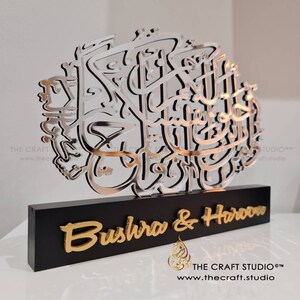 Op de afbeelding: Een zilveren en zwarte decoratieve wandkunst met de Arabische kalligrafie "Bismillah" en de namen "Bushra & Haroon" in gouden letters op een zwarte basis. Het kunstwerk is gemaakt door The Craft Studio.