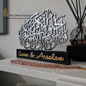 Op de afbeelding: Een zilveren gespiegelde kalligrafiekunstwerk met de tekst "Sana & Arsalan" op een zwarte basis. Het kunstwerk staat op een wit rek met een roze kralenketting op de voorgrond.