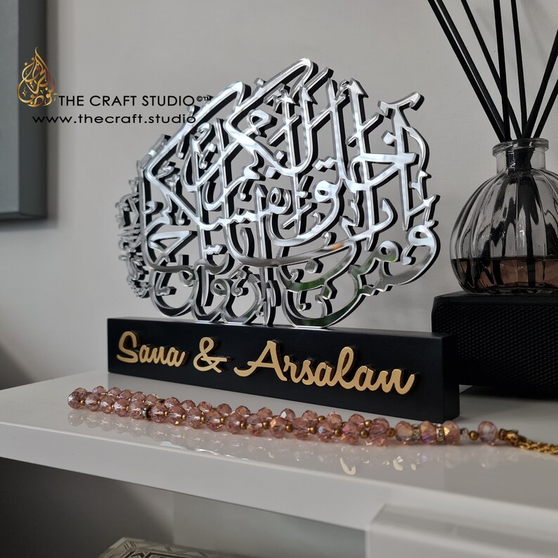 Op de afbeelding: Een zilveren gespiegelde kalligrafiekunstwerk met de tekst "Sana & Arsalan" op een zwarte basis. Het kunstwerk staat op een wit rek met een roze kralenketting op de voorgrond.