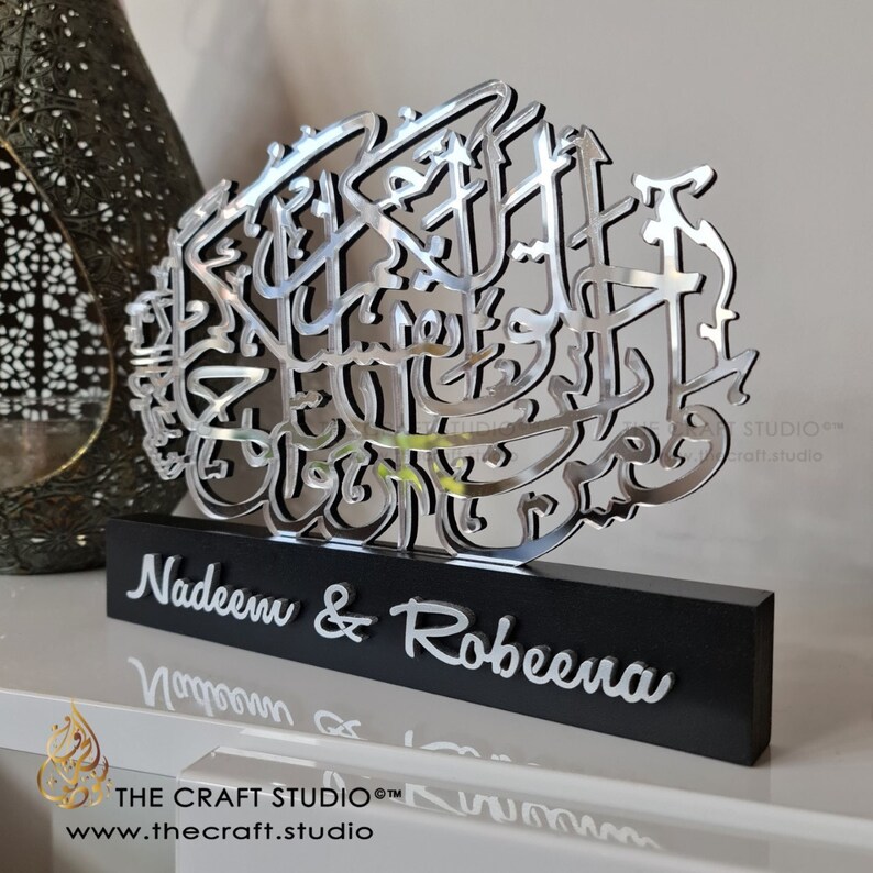 Op de afbeelding: Een zilveren metalen kalligrafiekunstwerk met de Arabische zin "Allah" in een decoratief lettertype. Het kunstwerk is gemonteerd op een zwarte houten basis met de namen "Nadeem & Robeena" gegraveerd in zilveren letters. De basis bevat ook de tekst "THE CRAFT STUDIO" en "www.thecraft.studio".