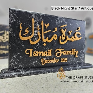 Peut inclure: Plaque noire effet marbre avec calligraphie arabe dor&eacute;e et texte anglais. Le texte indique "Umrah Mubarak", "Ismail Family" et "D&eacute;cembre 2023". La plaque a une base d&eacute;corative et est &eacute;tiquet&eacute;e "Black Night Star / Antique Gold".