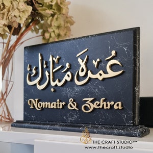 Peut inclure: Plaque rectangulaire en marbre noir avec des inscriptions dor&eacute;es. Le texte arabe est en haut, et les noms "Nomair & Zehra" sont en dessous. Le logo et le site web de The Craft Studio sont visibles. Fleurs s&eacute;ch&eacute;es en arri&egrave;re-plan.