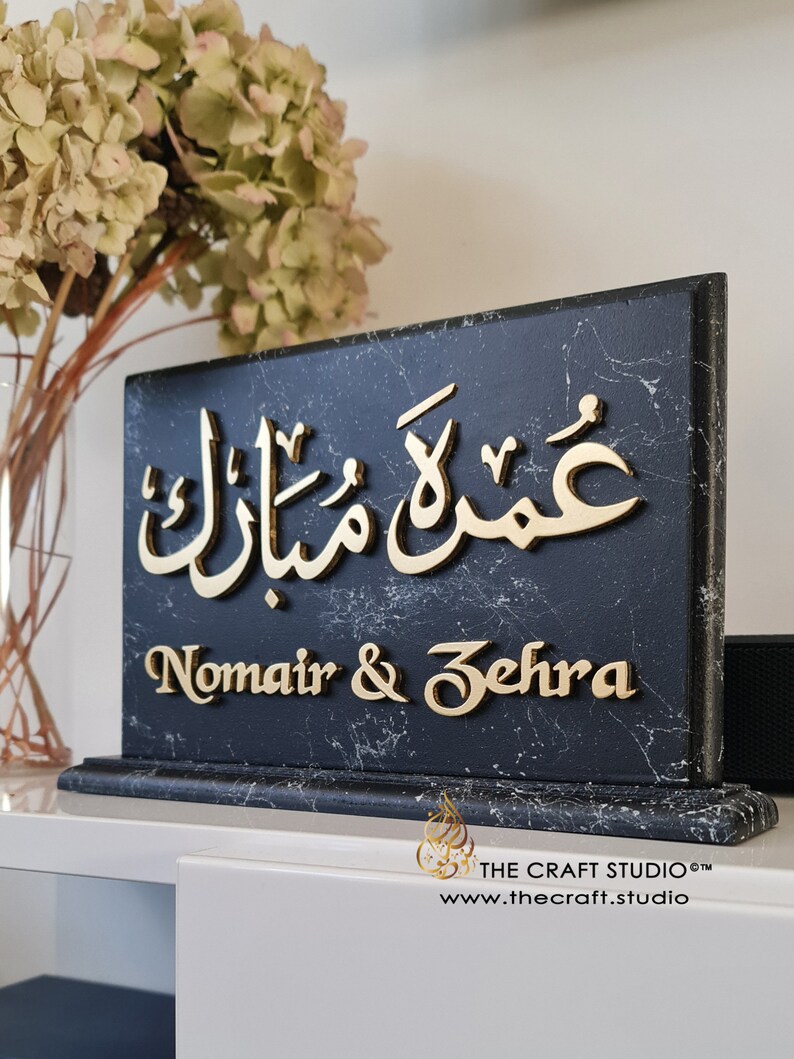 Peut inclure: Plaque rectangulaire en marbre noir avec des inscriptions dor&eacute;es. Le texte arabe est en haut, et les noms "Nomair & Zehra" sont en dessous. Le logo et le site web de The Craft Studio sont visibles. Fleurs s&eacute;ch&eacute;es en arri&egrave;re-plan.