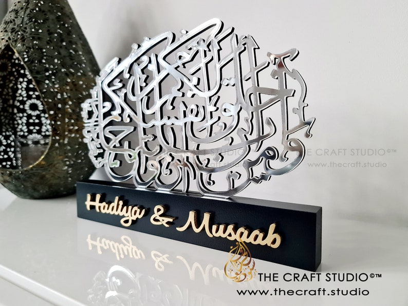 Op de afbeelding: Een zilveren metalen wandkunst met Arabische kalligrafie op een zwarte houten basis. De basis heeft de namen "Hadiya & Musaab" in gouden letters.
