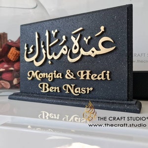 Peut inclure: Une plaque noire avec des lettres dorées. La plaque porte la phrase arabe "عمرة مبارك" et les noms "Mongia & Hedi Ben Nasr". La plaque est posée sur une surface blanche.