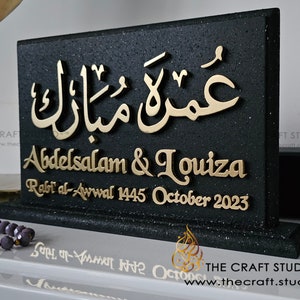 Peut inclure: Plaque noire d&eacute;corative avec des lettres dor&eacute;es. Le texte en arabe est sur le dessus, suivi des noms "Abdelsalam & Louiza" et la date "Octobre 2023". Le logo The Craft Studio est en bas.