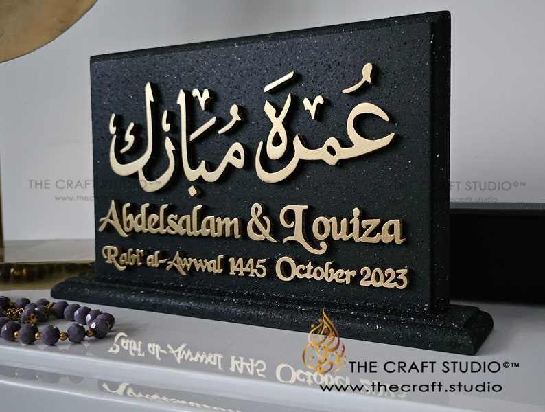 Peut inclure: Plaque noire d&eacute;corative avec des lettres dor&eacute;es. Le texte en arabe est sur le dessus, suivi des noms "Abdelsalam & Louiza" et la date "Octobre 2023". Le logo The Craft Studio est en bas.
