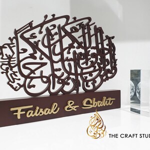 May include: A wooden decorative plaque with intricate Arabic calligraphy in brown. The plaque has a base with the names "Faisal & Shabdit" in gold lettering. The plaque is on a white surface with a clear glass block in the background.
