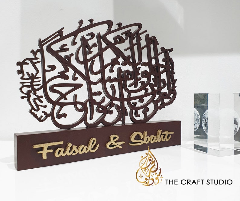 May include: A wooden decorative plaque with intricate Arabic calligraphy in brown. The plaque has a base with the names "Faisal & Shabdit" in gold lettering. The plaque is on a white surface with a clear glass block in the background.