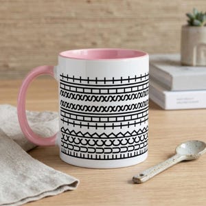 May include: White ceramic mug with a pink interior and handle. The mug has a black geometric pattern around the outside. A small ceramic spoon sits on the wooden surface beside the mug.