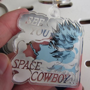 Puede incluir: Un llavero de acrílico transparente con un personaje de dibujos animados con cabello azul y un cigarrillo, con el texto "See You Space Cowboy..." en un globo de diálogo.