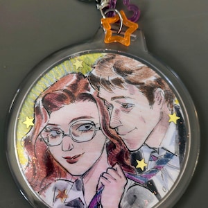 Puede incluir: Un llavero redondo con una ilustración en acuarela de una pareja. La mujer tiene el pelo rojo y gafas, tirando de la corbata del hombre. El llavero tiene un anillo plateado, un corazón morado y un charm de estrella naranja.
