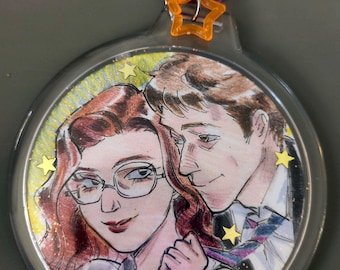 May include: A round keychain featuring a watercolor illustration of a couple. The woman has red hair, glasses, and is pulling on the man's tie. The keychain has a silver ring, a purple heart, and an orange star charm.