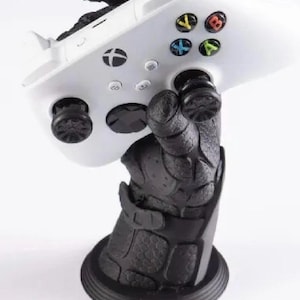 Könnte beinhalten: Ein weißer Xbox-Controller, der von einem schwarzen, strukturierten, handförmigen Ständer gehalten wird. Der Controller hat farbige Tasten und schwarze Joysticks. Der Ständer hat einen runden Sockel.